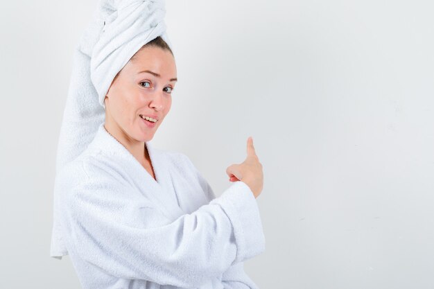 Young lady pointing back in white bathrobe, towel and looking merry. front view.