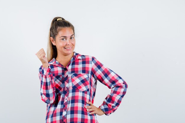 Young lady pointing back with thumb in checked shirt and looking cheerful. front view.