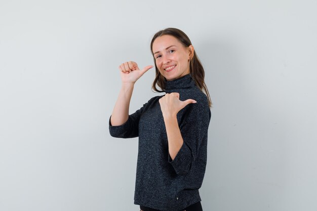 Young lady pointing back with thumbs in casual shirt and looking merry. front view.