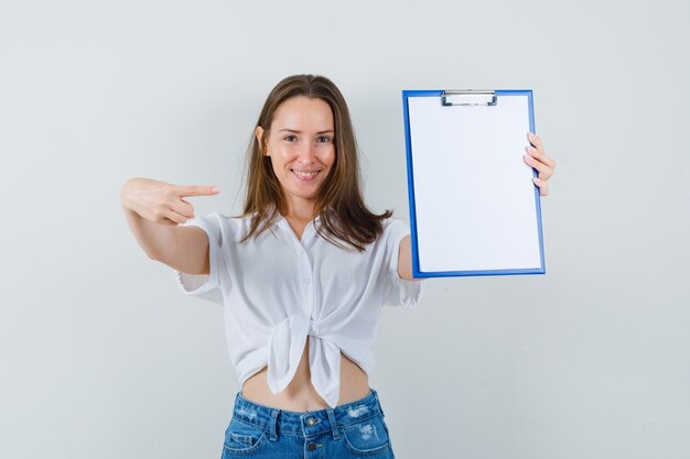 Young lady pointing to blank clipboard in white blouse front view.