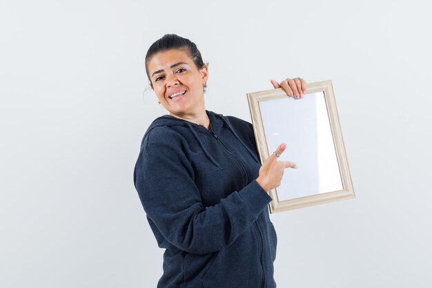 Young lady pointing at blank frame in jacket and looking glad. front view.