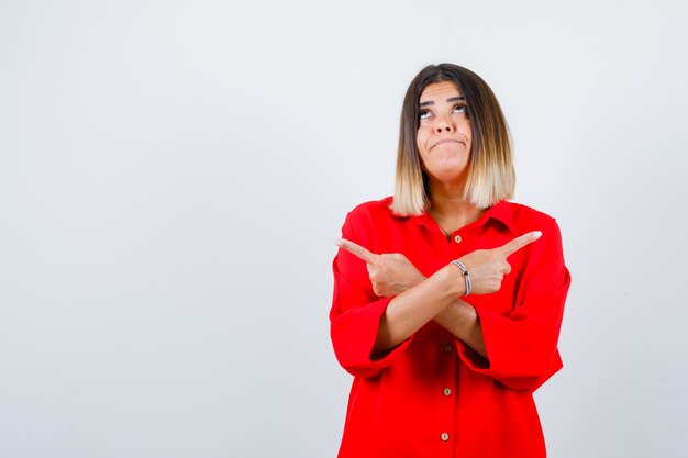 Young lady pointing to the both sides in red oversize shirt and looking indecisive , front view.