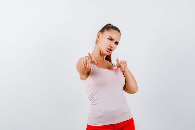 Young lady pointing at camera in beige tank top and looking cute. front view.