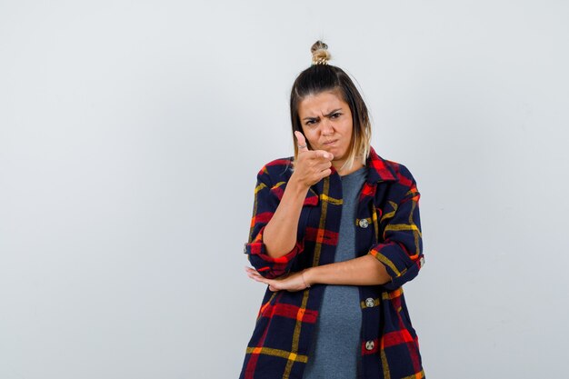 Young lady pointing at camera in casual checked shirt and looking dismal , front view.