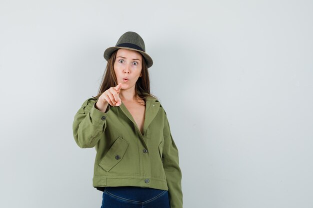 Молодая дама, указывая на камеру в куртке, штанах, шляпе и удивленно смотрит