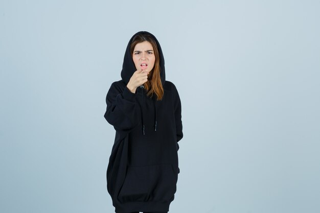 Young lady pointing camera in oversized hoodie, pants and looking angry. front view.