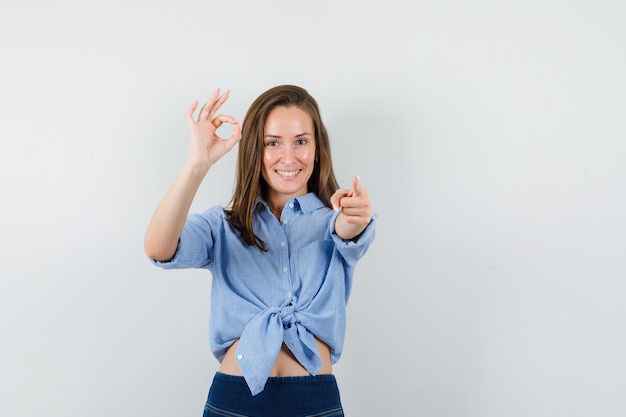 Young lady pointing at camera with ok sign in blue shirt, pants and looking glad