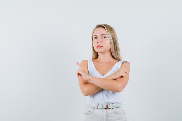 Young lady pointing at different sides with crossed arms in white blouse and looking confused