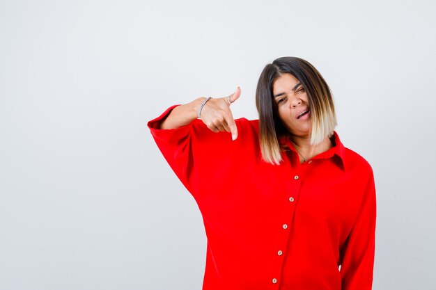 Young lady pointing down in red oversize shirt and looking self-confident. front view.