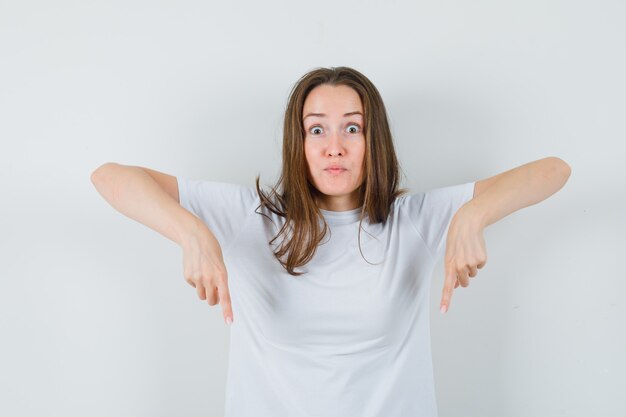 Young lady pointing down in white t-shirt and looking curious 