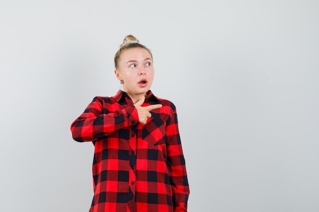 Young lady pointing at front in checked shirt and looking surprised