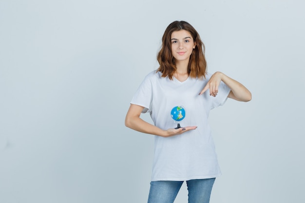 Tシャツ、ジーンズを保持し、自信を持って、正面図を見て地球を指している若い女性。