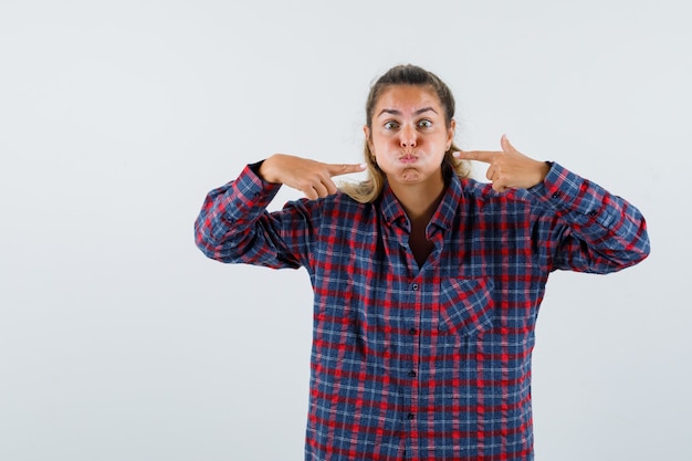 Foto gratuita giovane donna che indica le sue guance soffiate in camicia a quadri e sembra divertente, vista frontale.