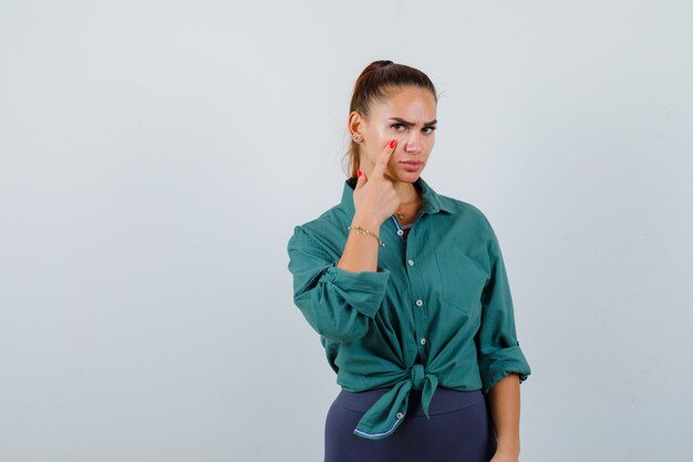 Young lady pointing at her eyelid in green shirt and looking upset. front view.