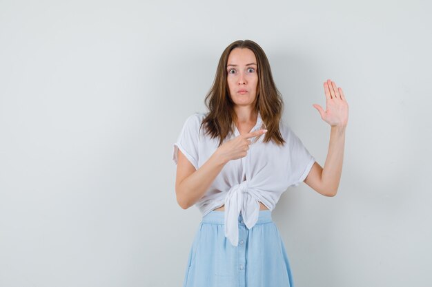 Young lady pointing at her palm in blouse, skirt and looking puzzled