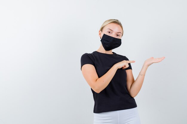 Young lady pointing at her palm spread aside in black t-shirt, mask and looking confident , front view.