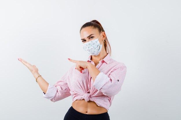 Young lady pointing at her palm spread aside in shirt, mask and looking confident , front view.