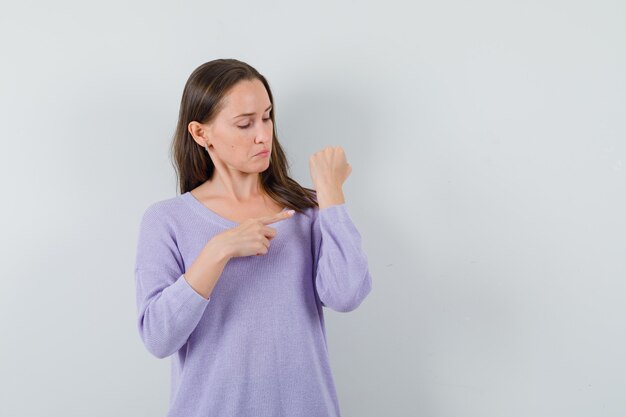 Young lady pointing at her wrist in casual shirt and looking confused 