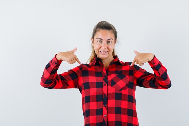 Young lady pointing at herself in checked shirt and looking proud