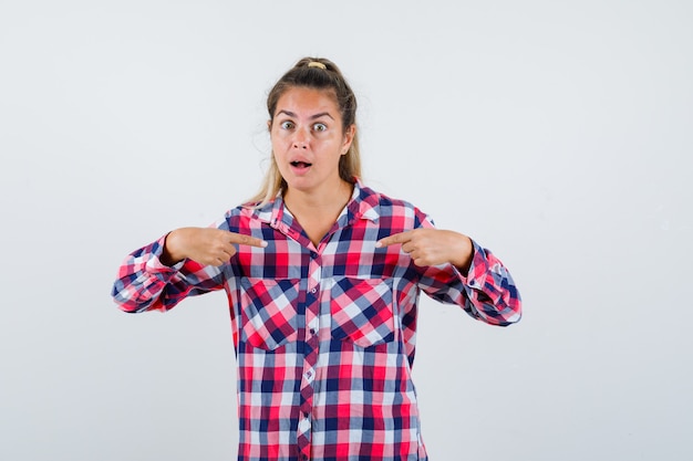 Young lady pointing at herself in checked shirt and looking puzzled. front view.