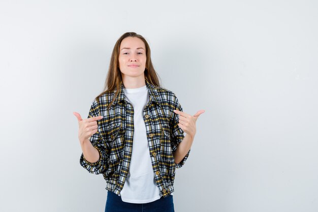 Tシャツ、ジャケット、ジーンズで自分を指差して誇らしげに見える若い女性。正面図。