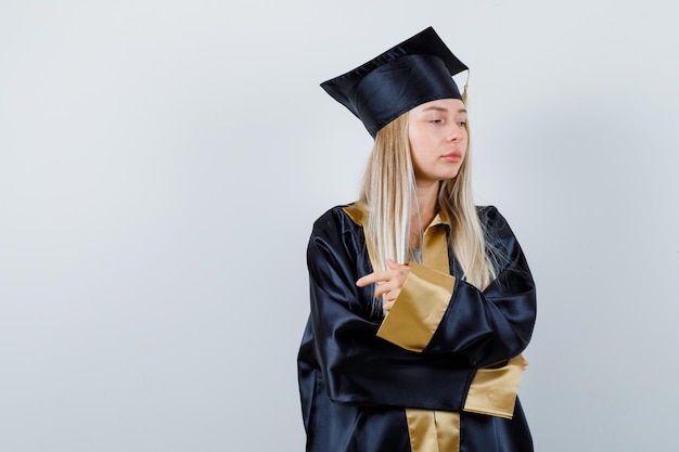 Young lady pointing to the left side in academic dress and looking thoughtful.