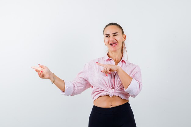 Giovane donna che punta a sinistra, sporge la lingua in camicia, pantaloni e sembra felice. vista frontale.