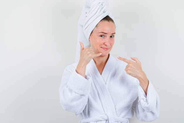 Young lady pointing at phone sign in white bathrobe, towel and looking pleased. front view.