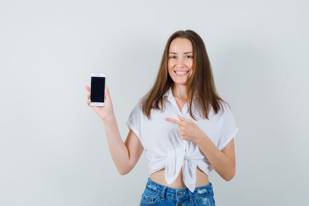 Young lady pointing at phone in white blouse and looking happy. front view.