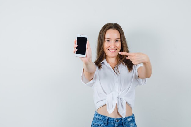 Young lady pointing at phone in white blouse and looking merry , front view.
