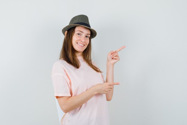 Young lady pointing to the right side in pink t-shirt, hat and looking jolly. front view.