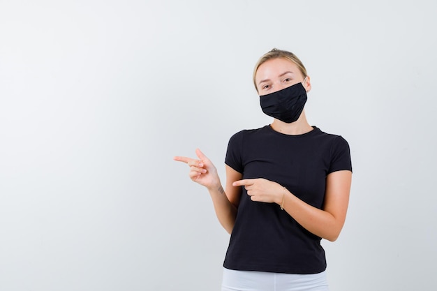 Young lady pointing to the right side in t-shirt, pants, medical mask and looking cheery