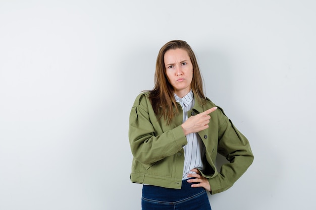 Young lady pointing to the right side while frowning in shirt, jacket and looking hesitant. front view.