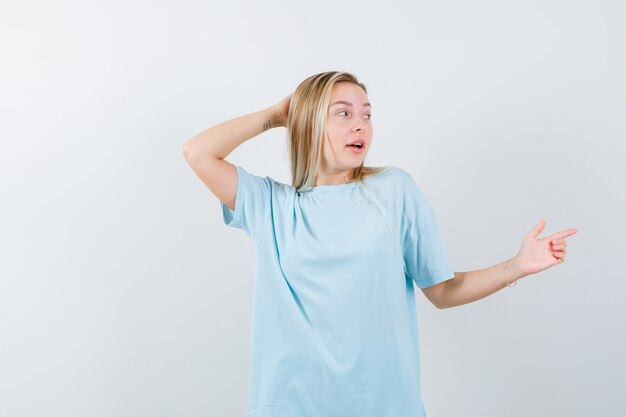 Young lady pointing to the right side while holding hand on head in t-shirt and looking puzzled , front view.