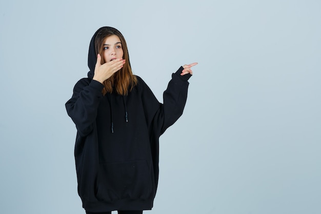 Foto gratuita giovane donna che indica il lato destro mentre tiene la mano sulla bocca in felpa con cappuccio oversize, pantaloni e sembra perplessa, vista frontale.