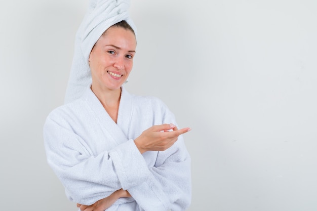 Young lady pointing to the right side in white bathrobe, towel and looking merry. front view.