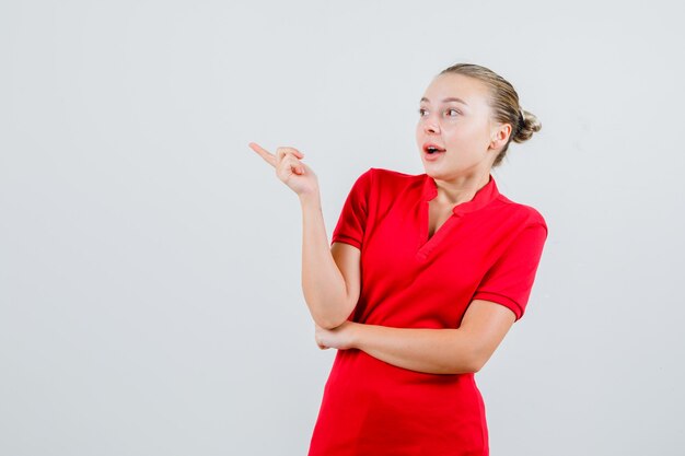 Young lady pointing to side in red t-shirt and looking astonished