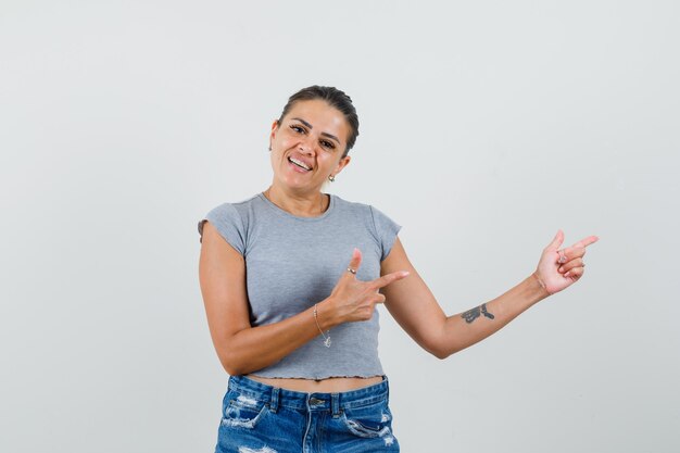 Young lady pointing to the side in t-shirt, shorts and looking cheerful. 