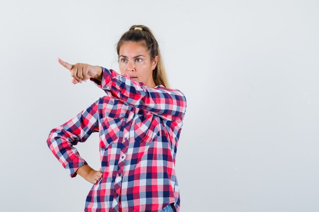 Young lady pointing at something away in checked shirt and looking angry. front view.