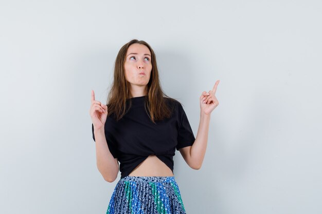 Young lady pointing up in blouse, skirt and looking hesitant