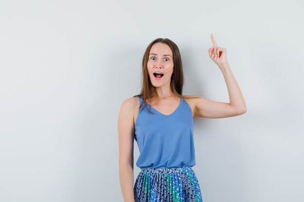 Young lady pointing up in blue blouse,skirt and looking amazed , front view. space for text