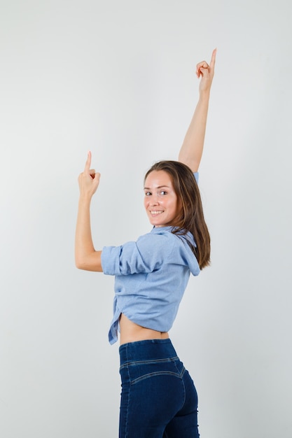 Молодая дама показывает вверх, протягивая руки в синей рубашке, штанах и выглядит весело
