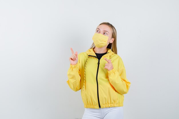 Young lady pointing up in jacket, mask and looking hopeful , front view.