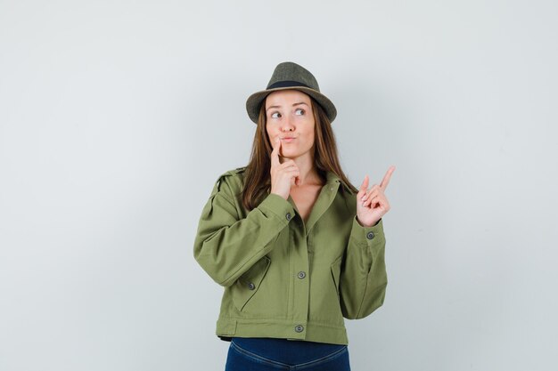 Young lady pointing up in jacket pants hat and looking pensive  