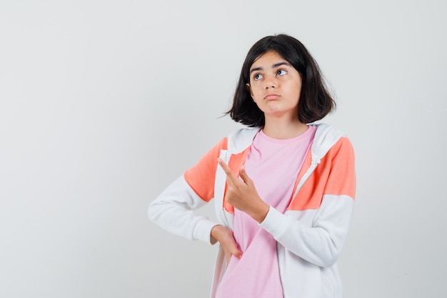 Young lady pointing up in jacket, pink shirt and looking thoughtful.