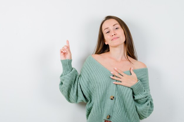 Young lady pointing up, keeping hand on chest in wool cardigan and looking proud. front view.