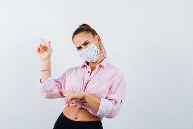 Young lady pointing up and left in shirt, mask and looking pensive , front view.