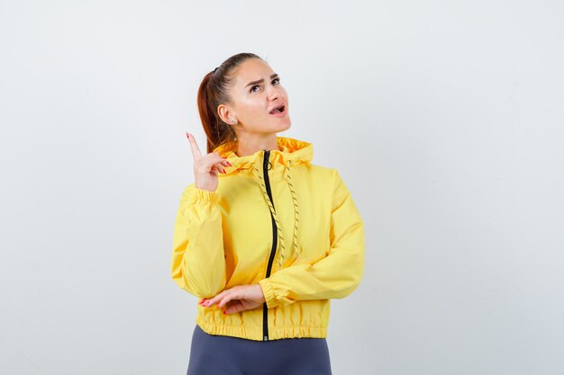 Young lady pointing up, opening mouth in yellow jacket and looking astonished . front view.