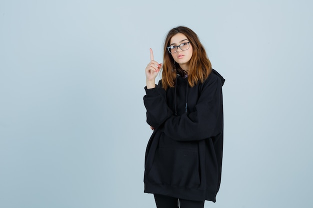 Young lady pointing up in oversized hoodie, pants and looking confident , front view.