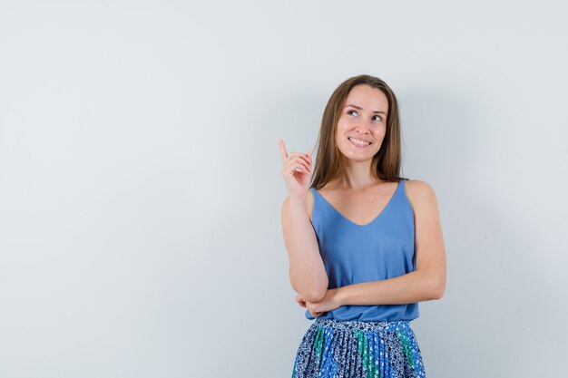 Young lady pointing up in singlet, skirt and looking cheerful. front view.